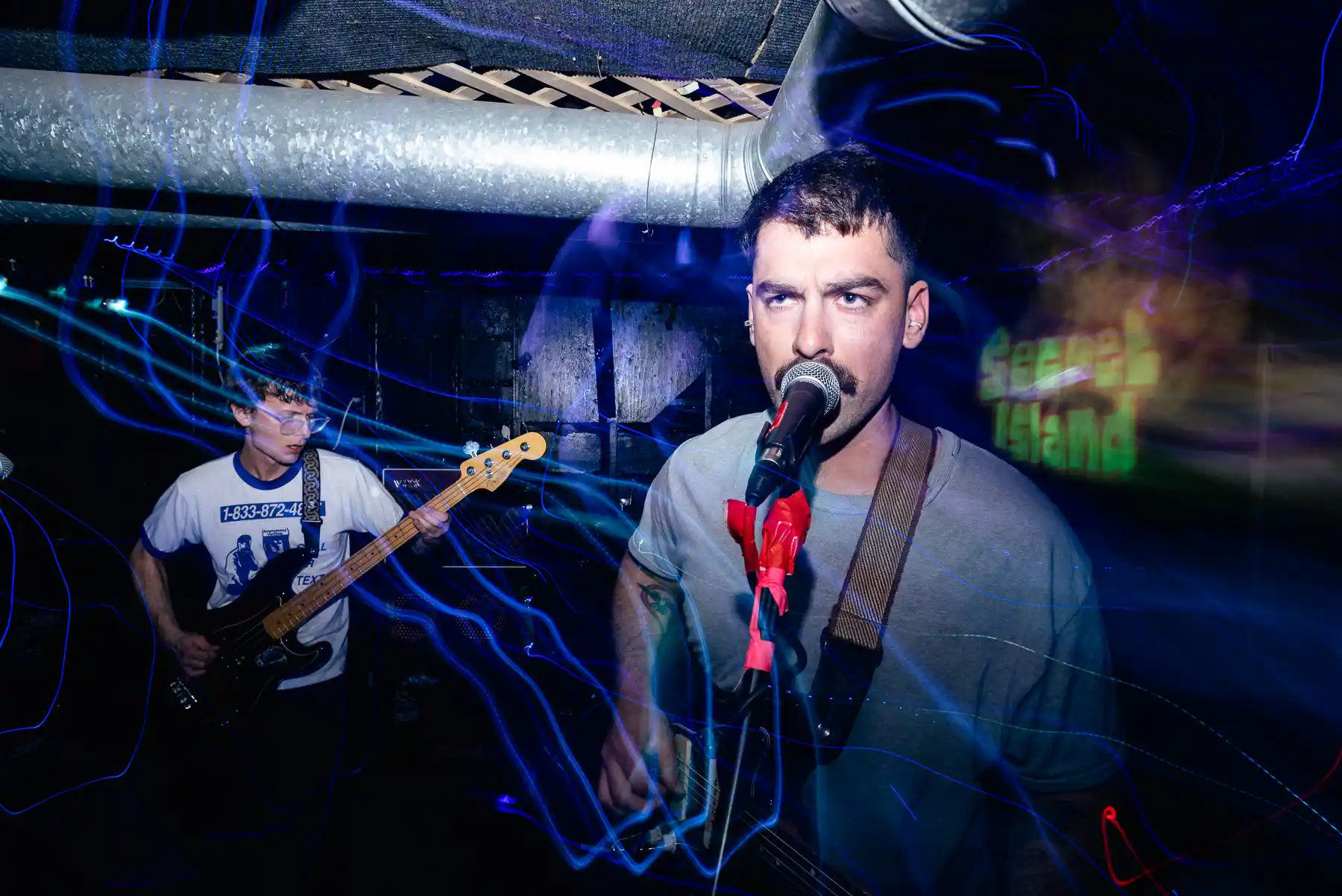 A man is singing into a microphone on stage while another man plays a guitar in the background, with blue swirling lights around them in a dimly lit setting.